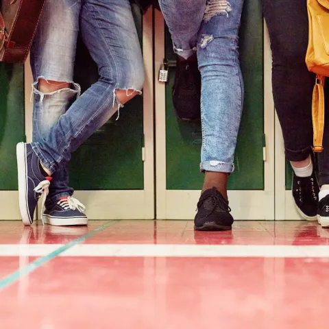 5 students standing in a row in front of lockers. Only legs and feet are shown