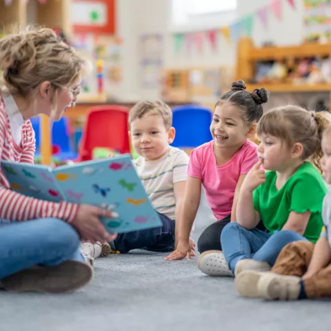 A preschool teacher sits on the floor of her classroom with a small group of students 