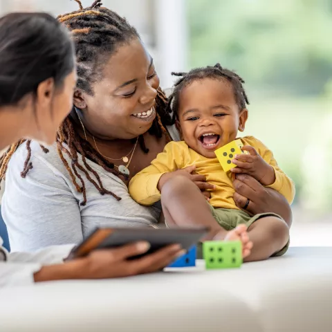 A young Mother brings in her toddler for a check-up. She is holding the little girl gently on the exam table as she talks with the doctor who is holding out a tablet with test results on it.