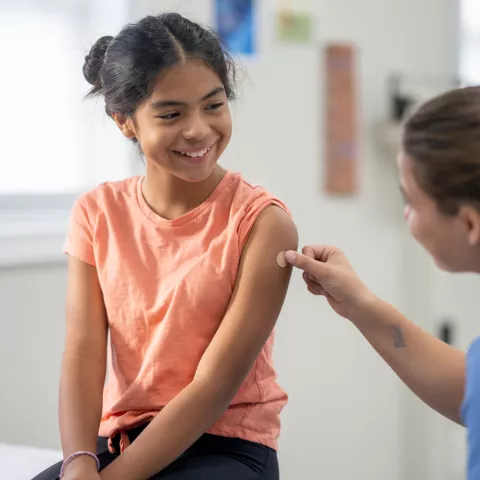 A school-aged girl sits up on an exam table as she holds still after a vaccination. She is dressed comfortably in a t-shirt and is smiling at her nurse as she places a bandage on the injection site.