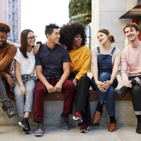 Six young adult friends sitting in a row on a bench in the street 
