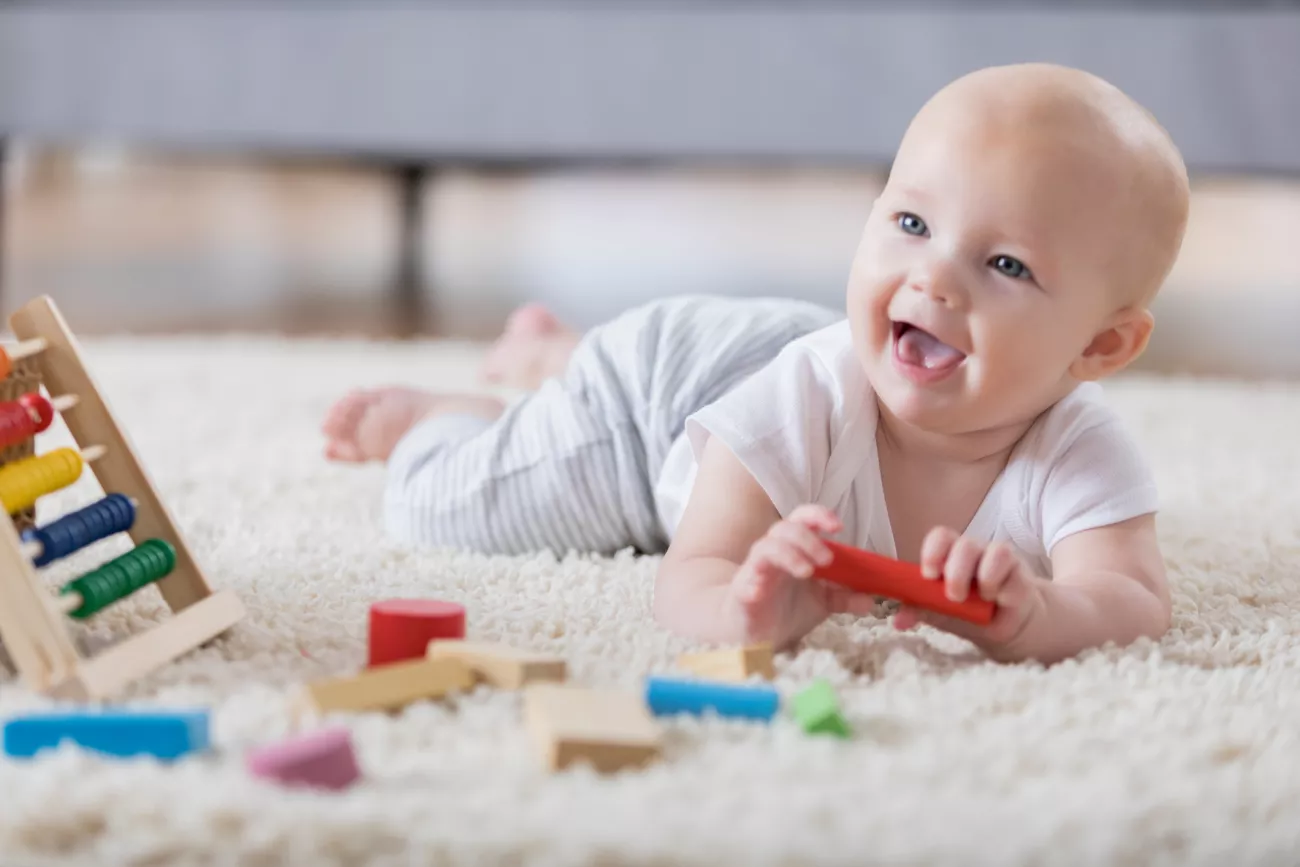 A smiling baby playing with toys on a carpet.
