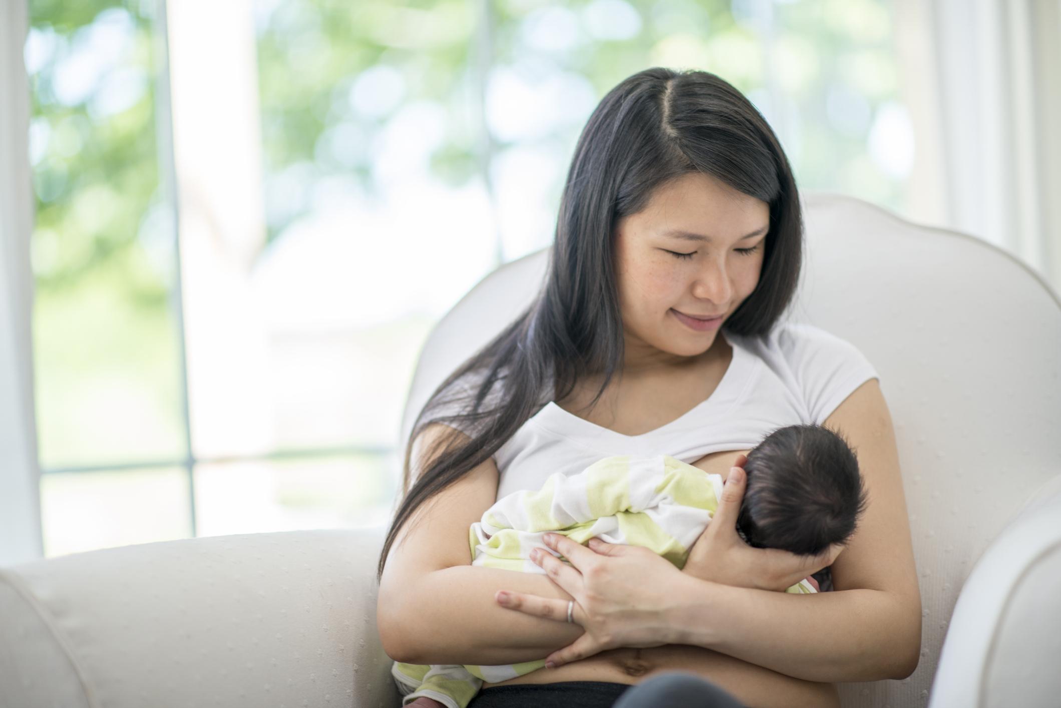 A mother and newborn baby boy are indoors in their house. The mother is breastfeeding while sitting in a chair.