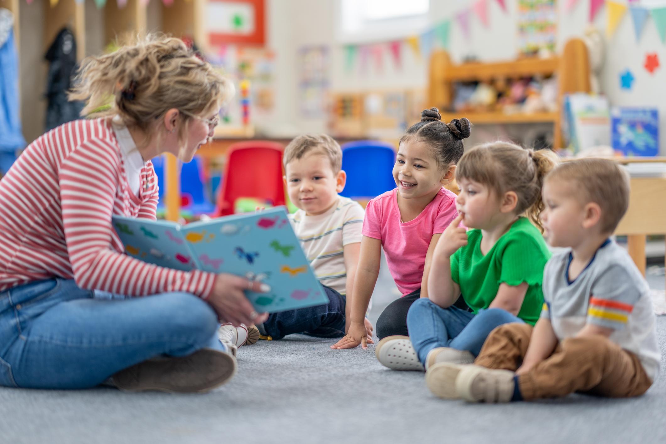 A preschool teacher sits on the floor of her classroom with a small group of students 