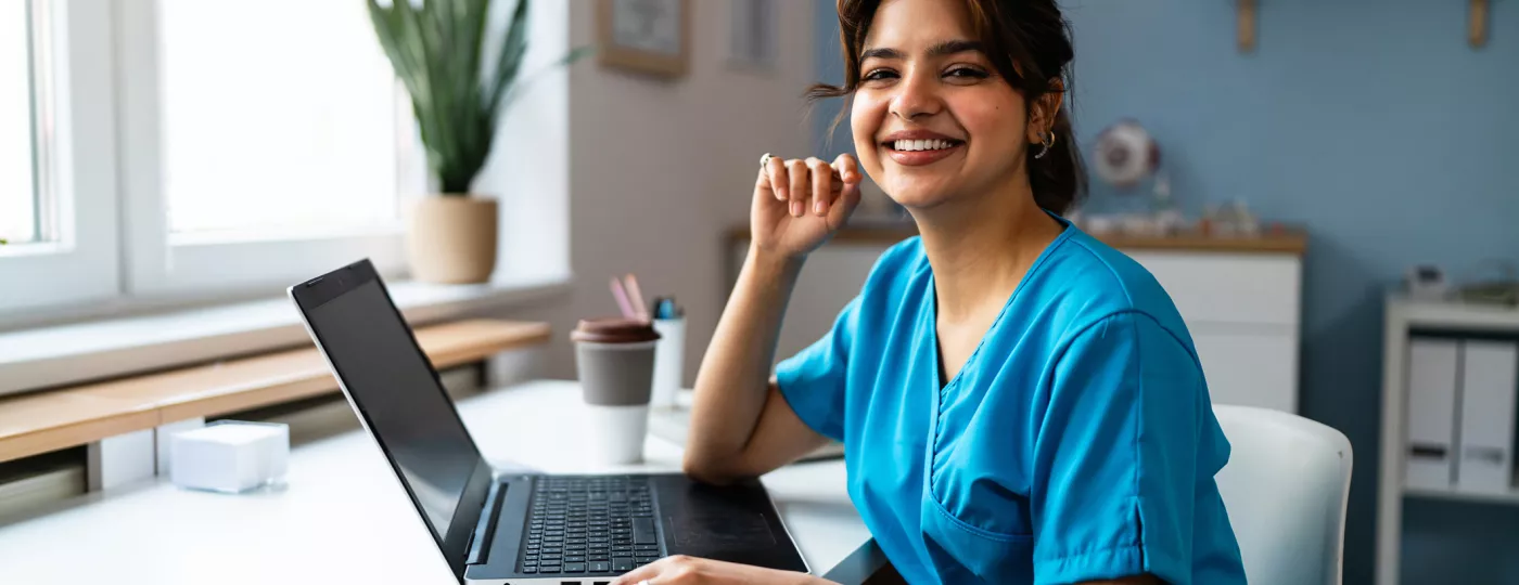 Portrait of a female doctor or a nurse of Indian ethnicity in a blue medical scrubs, using laptop at the modern ordination