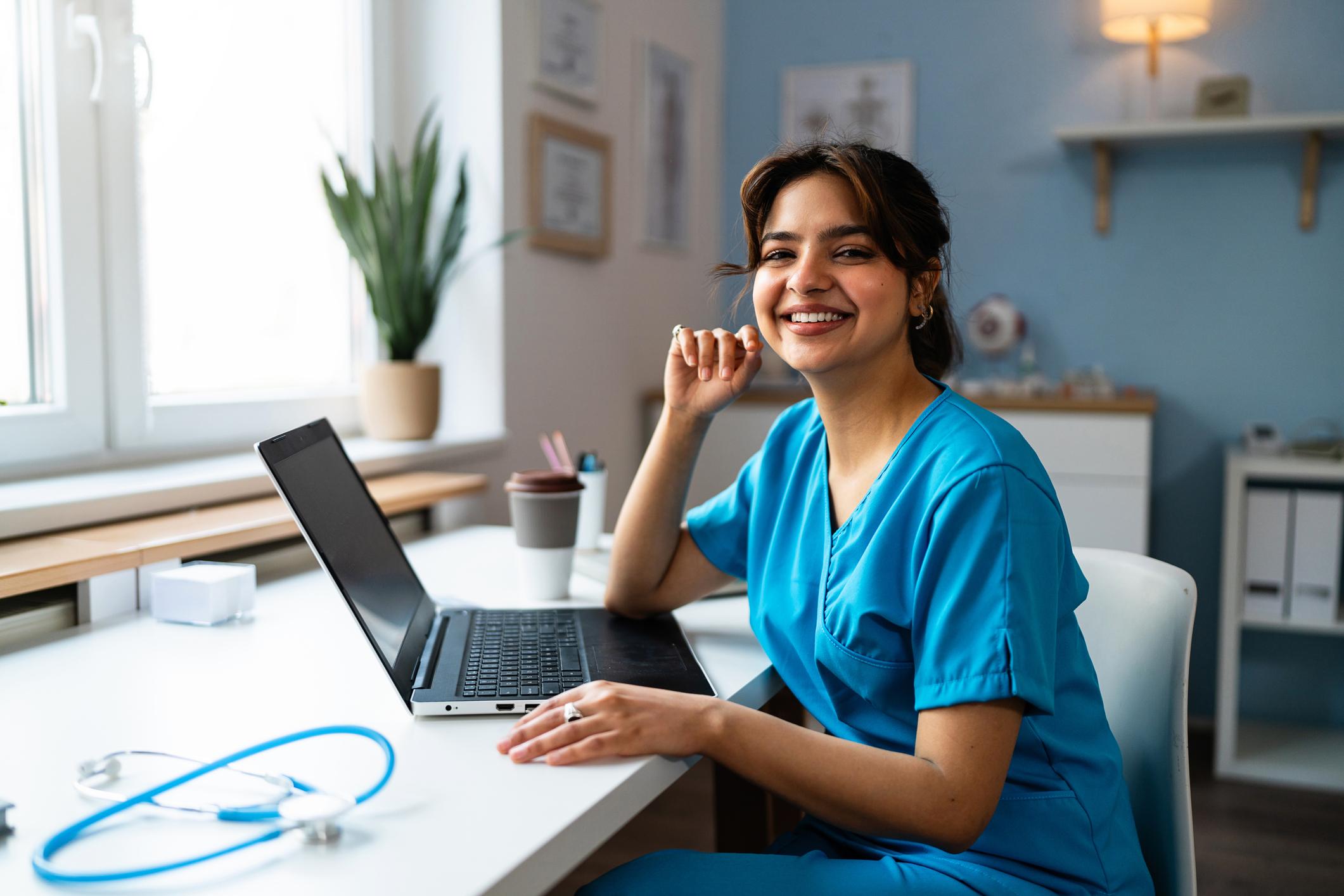 Portrait of a female doctor or a nurse of Indian ethnicity in a blue medical scrubs, using laptop at the modern ordination