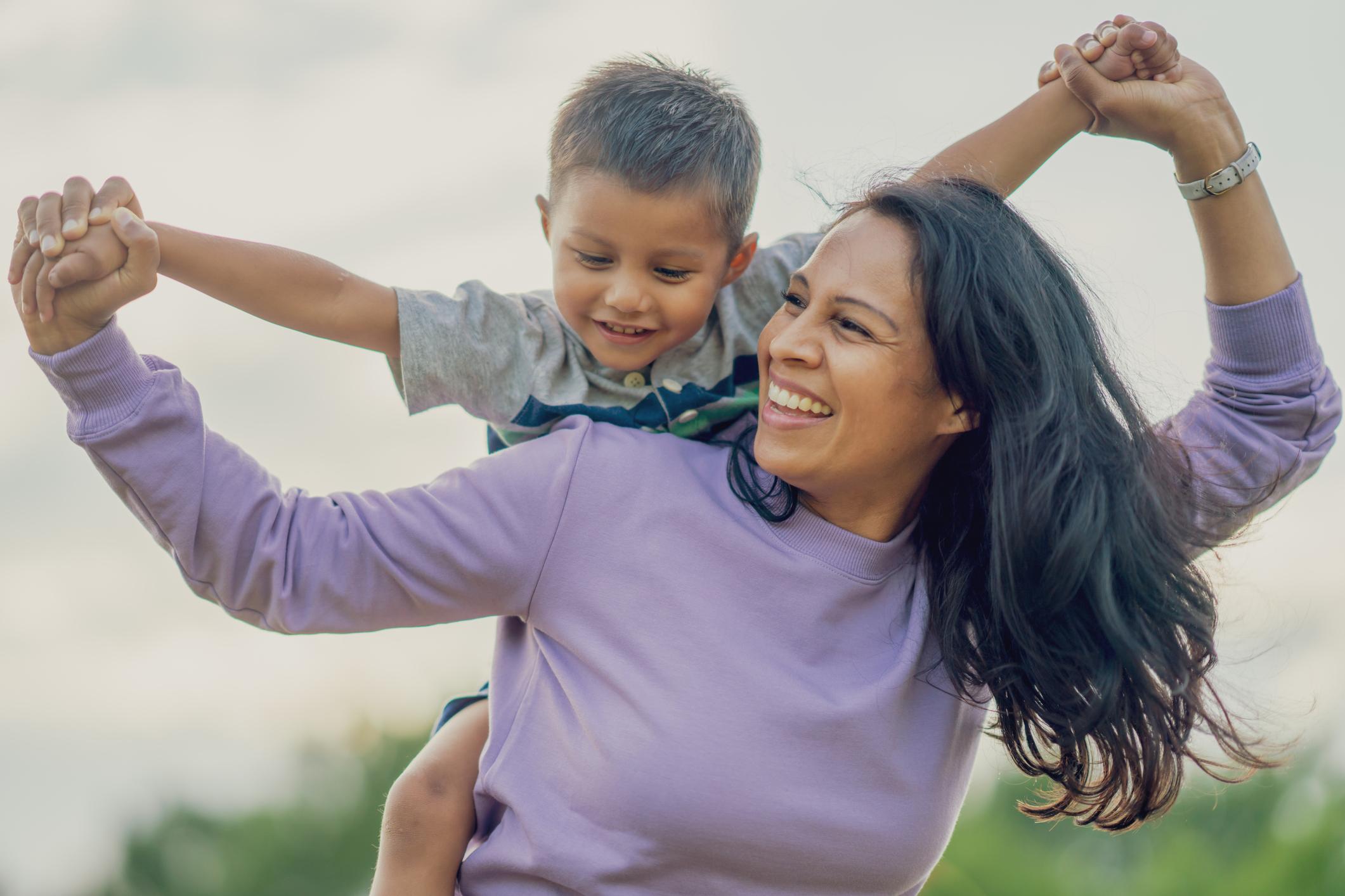A Mother holds her son on her back and his arms out wide, as she pretends to fly with him. They are both dressed casually and are smiling as they pose for a portrait outside.