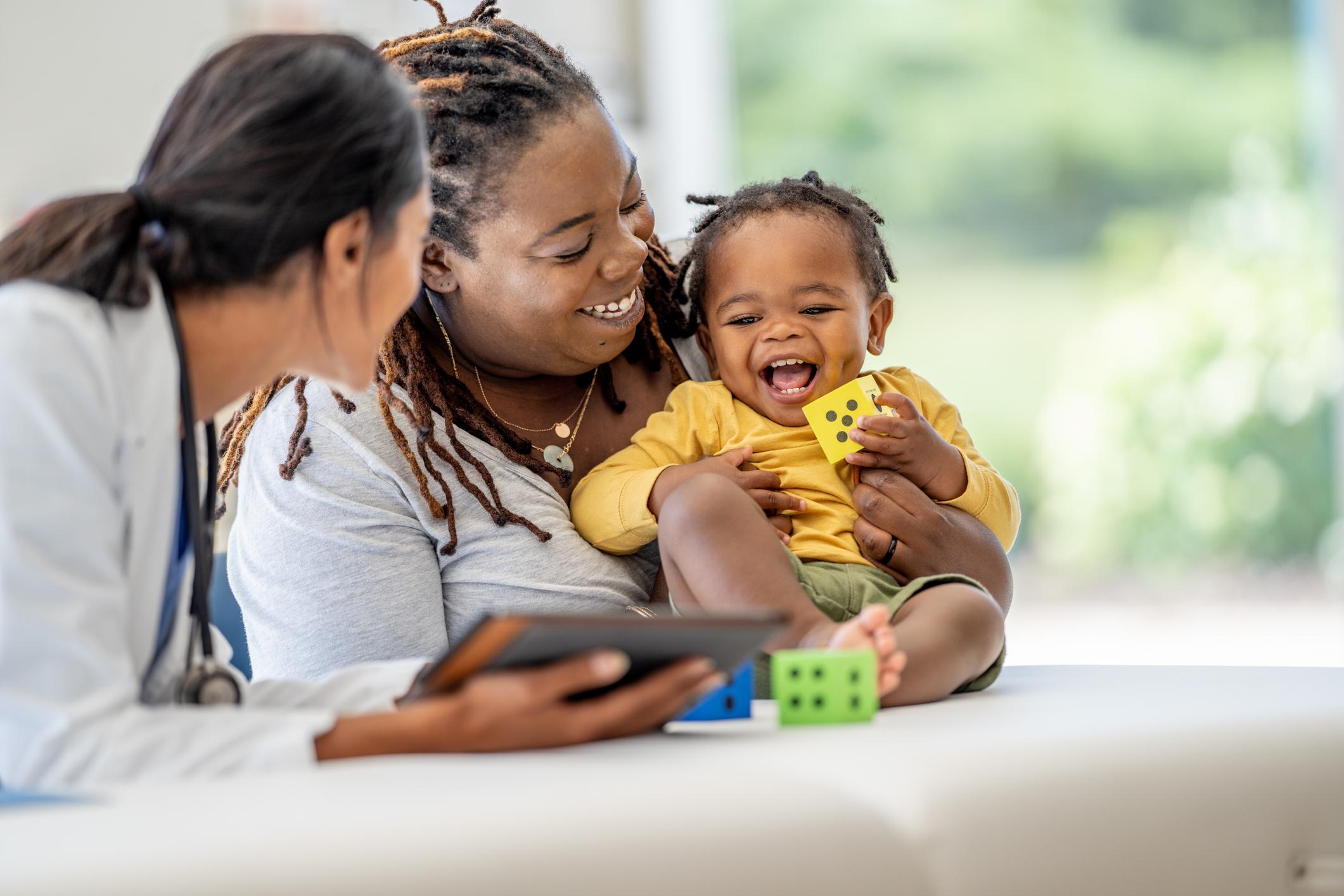A young Mother brings in her toddler for a check-up. She is holding the little girl gently on the exam table as she talks with the doctor who is holding out a tablet with test results on it.