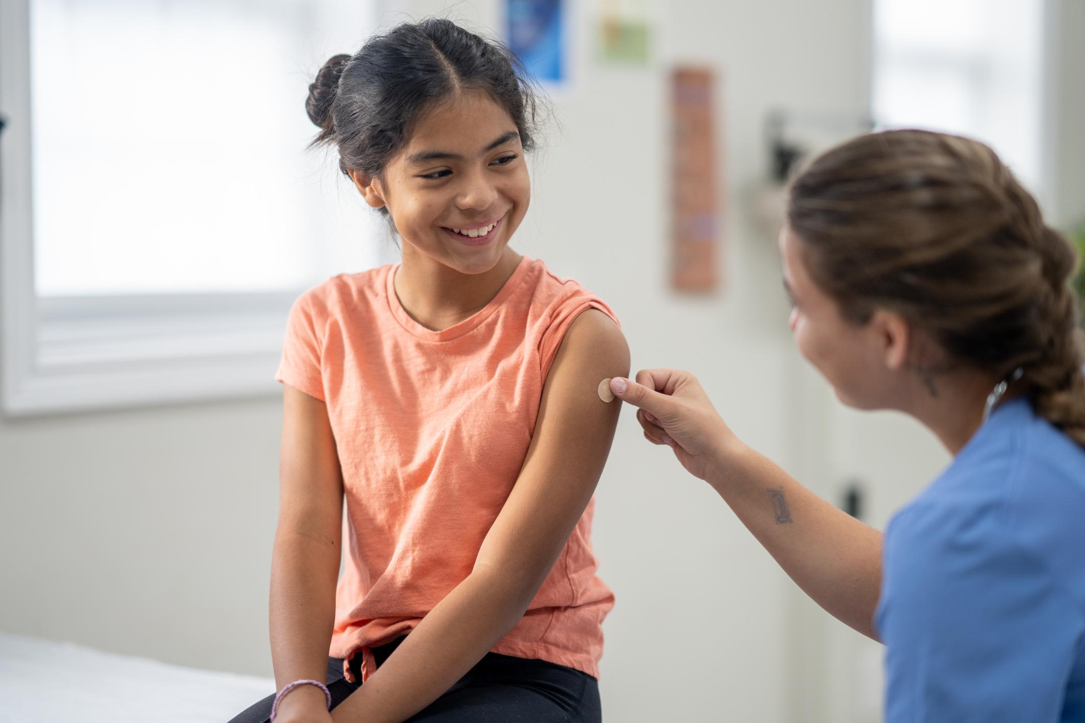 A school-aged girl sits up on an exam table as she holds still after a vaccination. She is dressed comfortably in a t-shirt and is smiling at her nurse as she places a bandage on the injection site.