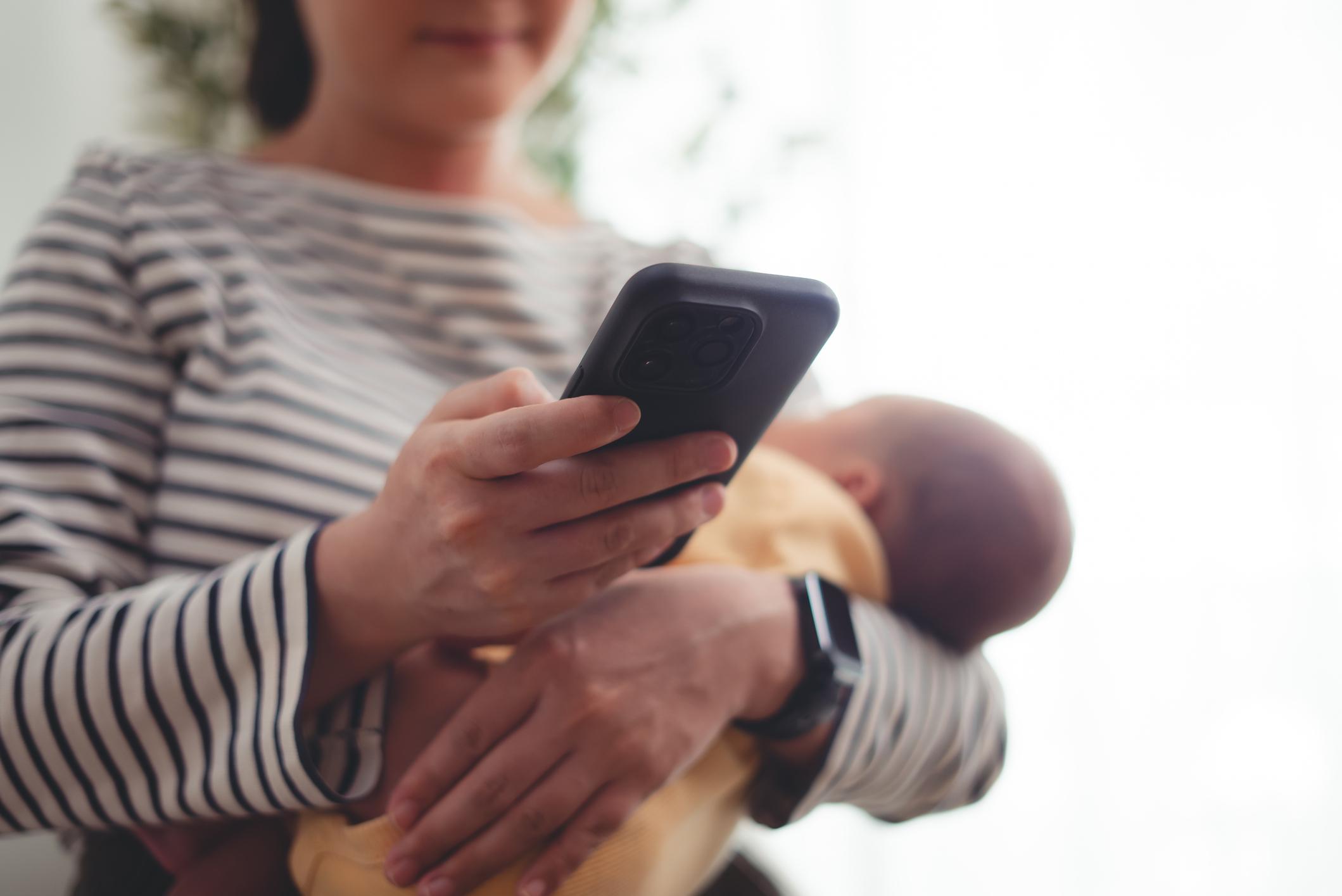 Close up shot of woman carrying her baby and taking a break using smartphone. 