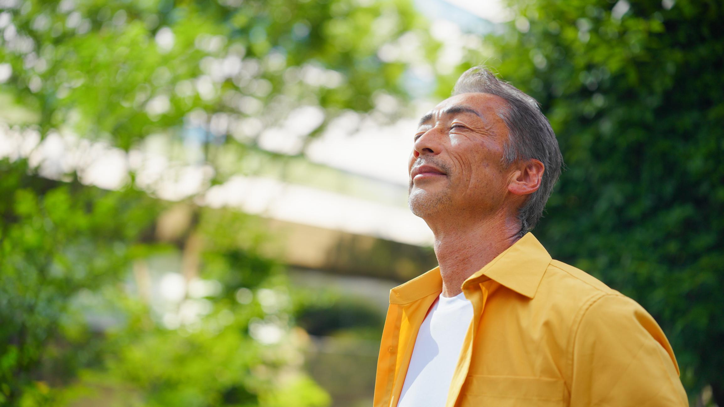A senior adult man is relaxing in the sun with his eyes closed in the city.