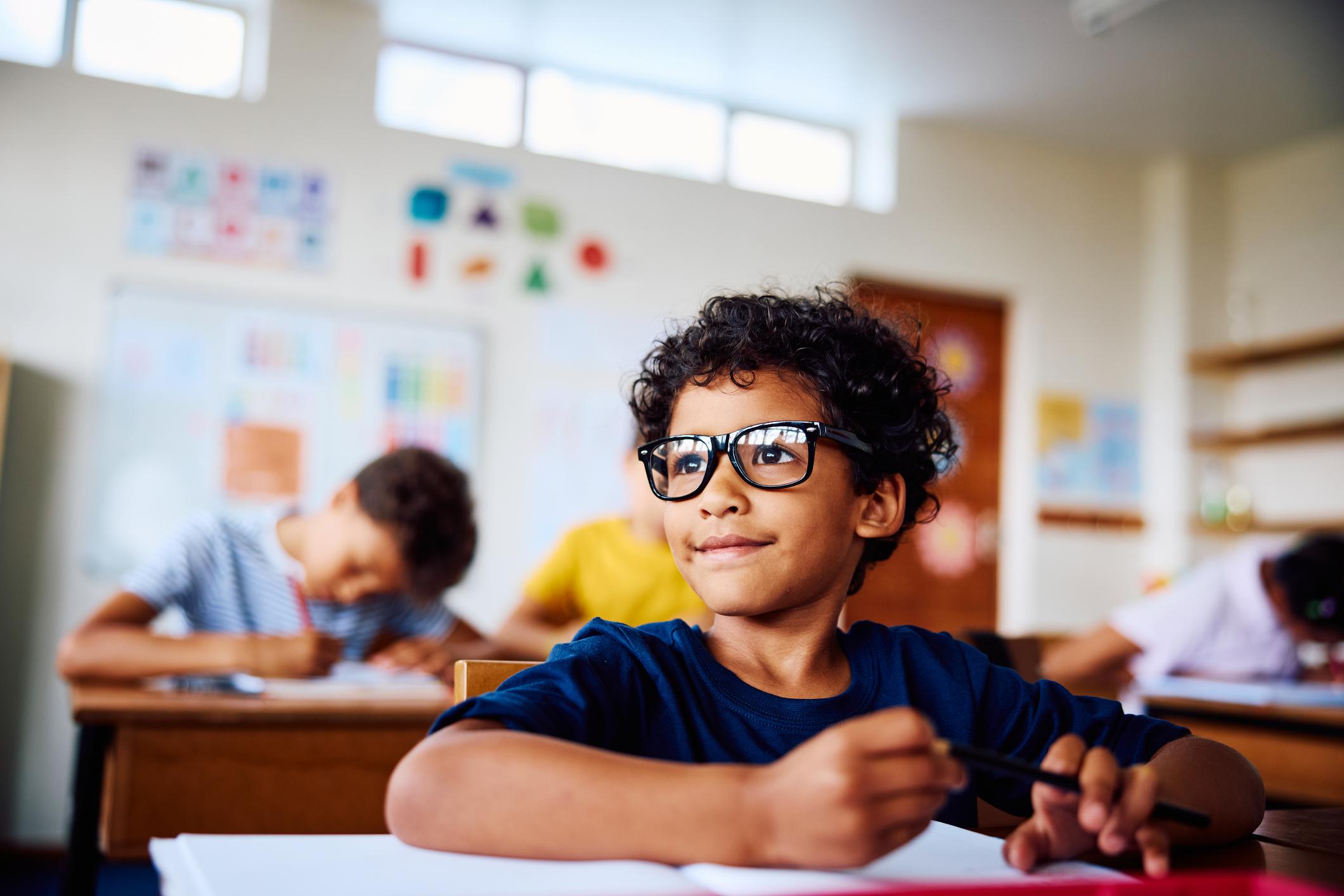 Schoolboy wearing eyeglasses sitting at desk, looking sideways, smiling in class