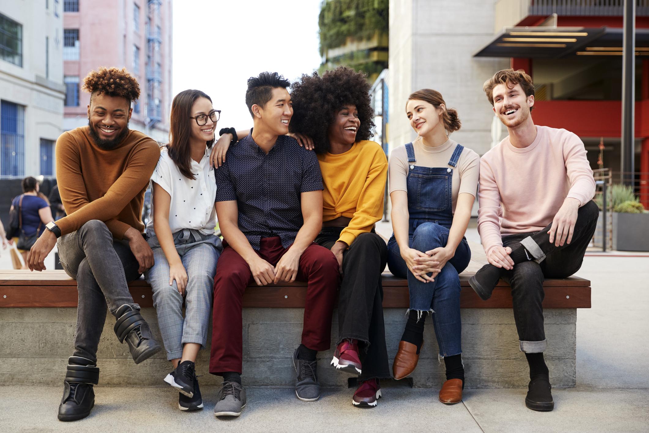 Six young adult friends sitting in a row on a bench in the street 