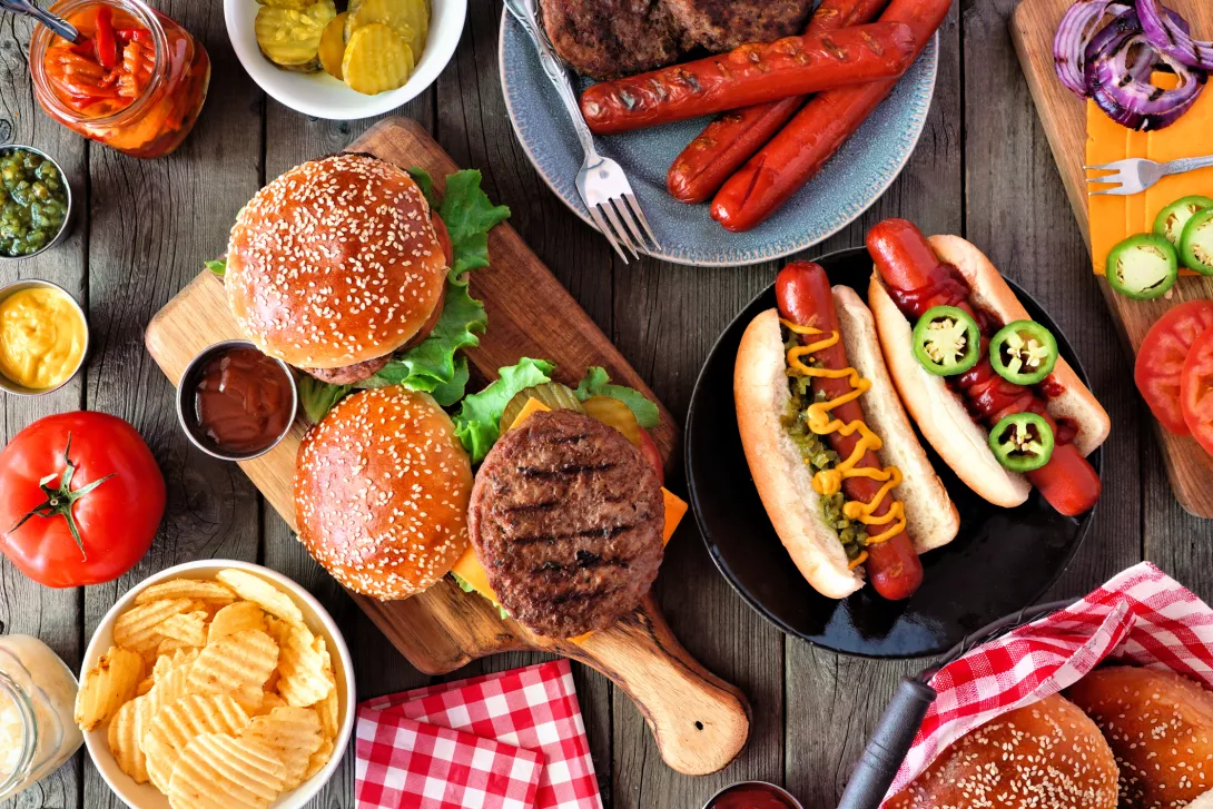 Summer BBQ food table scene with hot dog and hamburger buffet, top view over dark wood