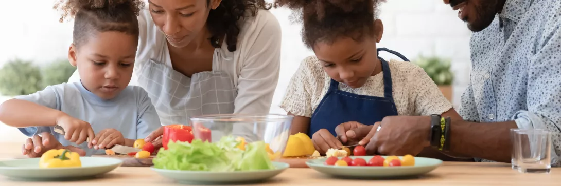 Happy family of four in the kitchen preparing food together. Parents are showing the two young sibling to slice fresh vegetables, peppers, tomatoes for salad on kitchen table or counter.