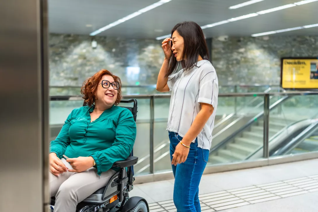 Smiling woman in a wheelchair and support worker talking waiting for elevator.