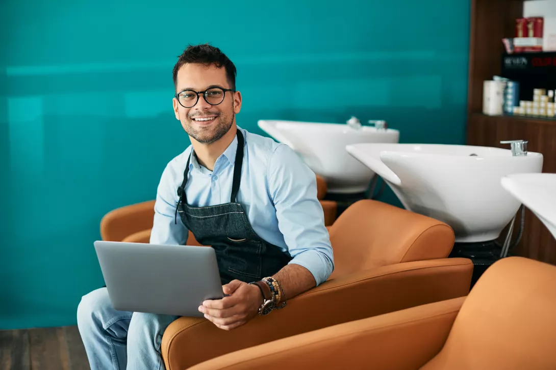 Young hairstylist using laptop while working in salon and looking at camera. 
