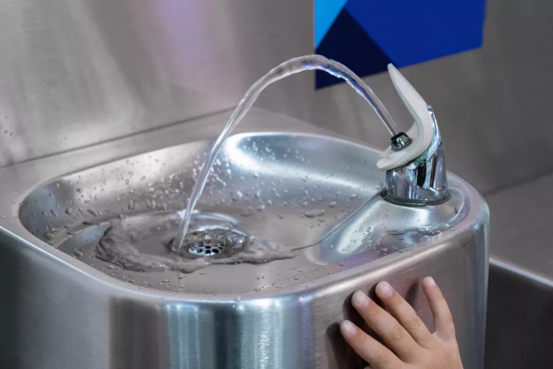 A child touches the handle of a modern drinking fountain as water flows in an indoor setting